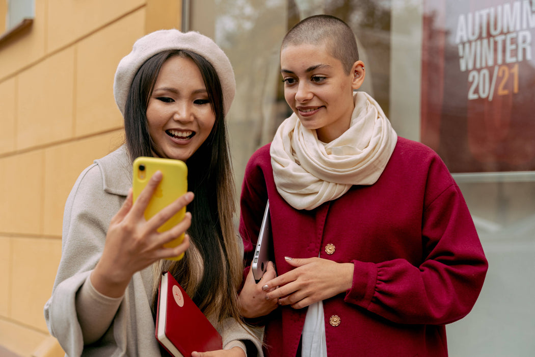 Two young women standing in an outdoor city space looking at a phone being held up by one of the women.